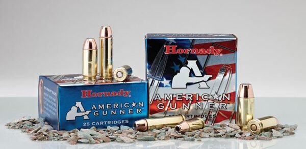 Boxes of hornady american gunner ammunition with cartridges standing and lying on scattered pebbles.