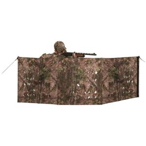 A person wearing camo is positioned behind a camouflage hunting blind, aiming a black firearm. The blind features a leafy green and brown pattern.