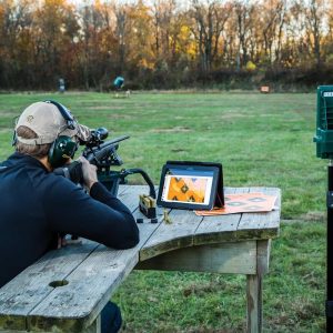 Man in a tan hat and green earmuffs aiming a rifle on a wooden bench, with a digital target display and ammo boxes beside him, set on a grassy field with autumn trees in the background.