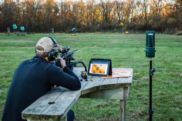 Man in a tan hat and green earmuffs aiming a rifle on a wooden bench, with a digital target display and ammo boxes beside him, set on a grassy field with autumn trees in the background.